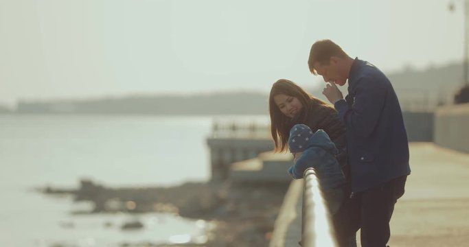 Happy international family stands at the parapet on the pier in spring. Korean woman walks with her husband and young son in the park. Circular motion of the camera.