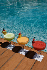 Red yellow and green cocktails on a wooden deck next to a pool