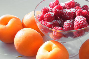 Raspberries ripe in a glass ice-cream bowl, orange apricots on the table on a white background