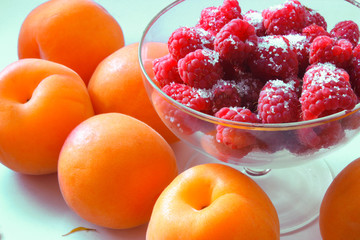 Raspberries ripe in a glass ice-cream bowl, orange apricots on the table on a white background