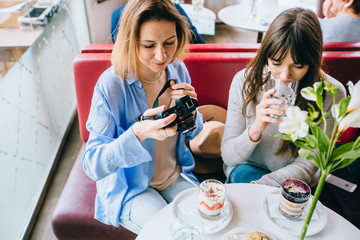 Two adult friends, sisters, attractive women eat dessert in a restaurant, cafeteria, drink from glass water, happy girls, brunette and blond, viewing photos on camera. Lifestyle travel concept.