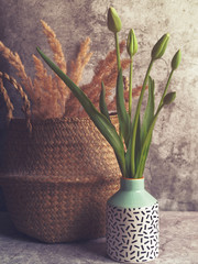 Five fresh tulips with closed buds are in a motley vase on a gray stone background. Behind is a woven straw basket with dried ornamental grass.