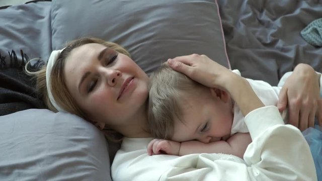 Young beautiful blonde mother lies on bed with little cute daughter, kisses and smiles at baby. Children bedtime