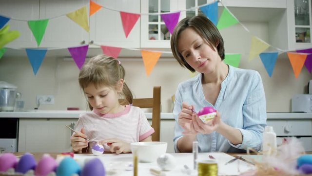 mother and daughter sitting at the table and crafting nest with colored eggs together and working in the kitchen