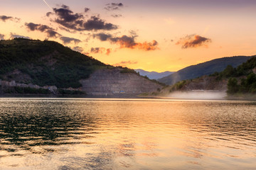 Golden sky and dramatic fluffy  clouds over reflective, silky lake and dust cloud flying over the water from the road