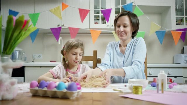 mother and daughter sitting at the table and crafting nest with colored Easter eggs together and luaghing on the kitchen