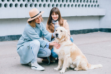 Two stylish women plays with a dog at city street urbam background. Training Golden retriever, the performance of the teams.