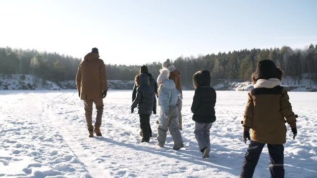 Three shot sequence of doting young couple with five kids enjoying winter hike on sunny day over snowy frozen lake, walking in line together, then children running race while parents watch and laugh