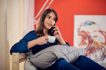Beautiful Caucasian brunette in pajamas sitting on the bed in bedroom, talking on the phone and...