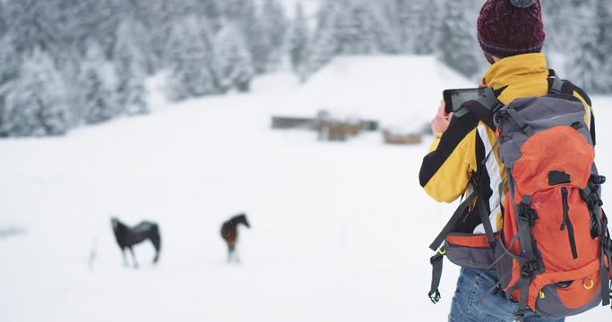 Young guy tourist arrived in amazing place in the middle of mountain , in the countryside he take pictures of horse , he wearing winter clothes and orange tourist bag