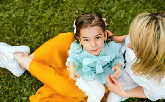 Top view image of happy kid playing with her mother, enjoying the time together outdoors. Cheerful little girl eating cotton candy with her mom, sitting on the green grass in the park. Mothers day.