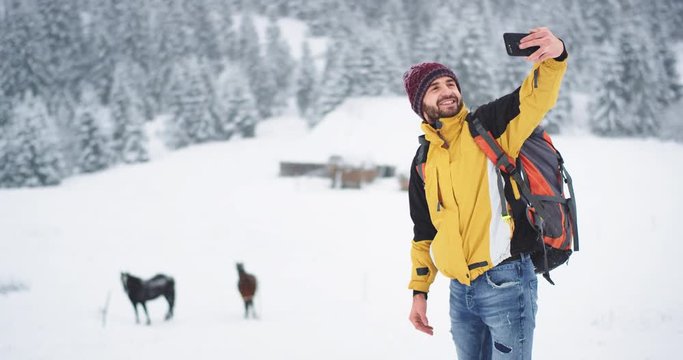 In the middle of winter tourist take pictures in mountain with two beautiful horses on the background , he have all travel equipment , happy smiling man take selfies with horses