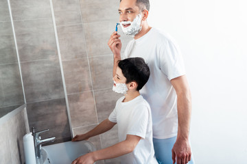 son with shaving cream on face near father shaving with razor in bathroom