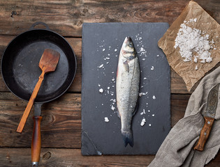 fresh whole sea bass fish on a black graphite board, next to it is an empty round black frying pan