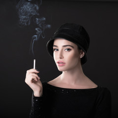 Obraz premium Young fashion woman smoking cigarette in hat and black drees over dark background. Vintage female portrait, styling. Image toned.