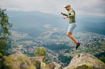 Lifestyle summer portrait of sportive man jumping  from stone on top of mountaing with beautiful landscape in front. Male traveler having fun among hils and rocks at wild nature outdoor.