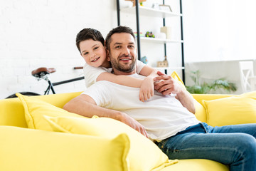 son looking at camera while hugging father sitting on couch at home