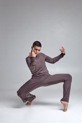 Photo of an athletic man ballet dancer dressed in a gray tracksuit, making a dance element against a gray background in studio.