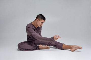 Photo of an athletic man ballet dancer dressed in a gray tracksuit, making a dance element against a gray background in studio.