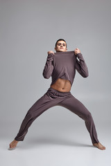 Photo of an athletic man ballet dancer dressed in a gray tracksuit, making a dance element against a gray background in studio.