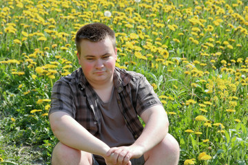 Naklejka na ściany i meble A young plump man sits in a meadow with blooming dandelions. Pensively looking in front of him.