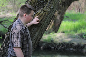 A young plump man put his hand on a tree trunk. Standing on the banks of the river watching the water. In the park among the bright green foliage.