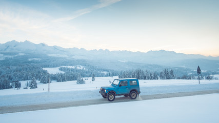 Niebieska klasyczna terenówka na ośnieżonej asfaltowej drodze z wysokimi zaspami śniegu, w tle pasmo górskie tatr i zachód słońca. © Tomasz