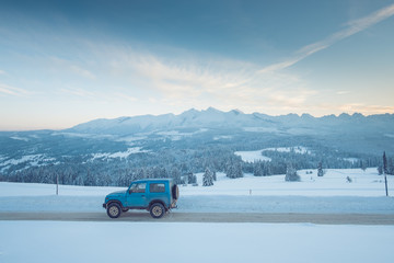 Niebieska klasyczna terenówka na ośnieżonej asfaltowej drodze z wysokimi zaspami śniegu, w tle pasmo górskie tatr i zachód słońca. © Tomasz