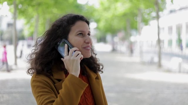Smiling young woman talking by cell phone outdoor. Beautiful happy woman with curly hair standing on street and having pleasant phone talk, handheld shot. Communication concept