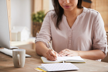 Mid section portrait of mixed race young woman writing notes in notebook while sitting at desk in office, copy space - Powered by Adobe