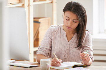 Portrait of beautiful Mixed race businesswoman writing in notebook while sitting at desk in office, copy space