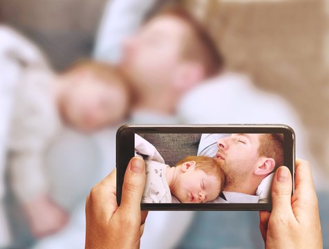 Young father sleeping with cute sleeping newborn baby lying on his chest with arms down. Mother is documenting the scene with a smartphone. Father and son blurred outside the mobile screen.