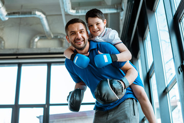 happy father giving piggyback ride to happy son in boxing gloves at gym