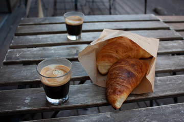 Zbliżenie na dwa kieliszki espresso i croissanty, stojące na drewnianym rustykalnym stoliku na zewnątrz kawiarni - obrazy, fototapety, plakaty