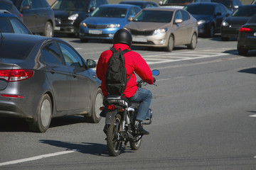 a man with a red jacket with a black backpack riding a moped under the road in heavy traffic
