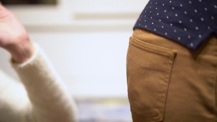 Female hand in a white pullover with a manicure slaps the guy's ass in brown pants and a dark blue shirt with white polka dots on a blurred light background in slow motion close-up.