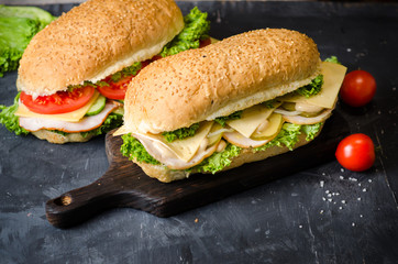 Two delicious sandwiches with ham, cheese, tomatoes, salty cucumber and salad over a wooden board on black background.