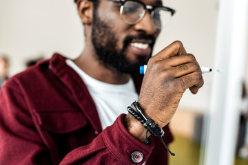 Selective focus of smiling African American student in glasses writing on flipchart