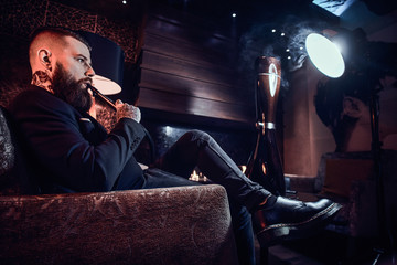 Handsome tattooed man is relaxing on the armchair and is smoking hookah.