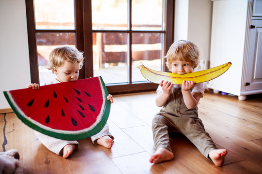 Two toddler children playing with large toy fruit indoors at home.
