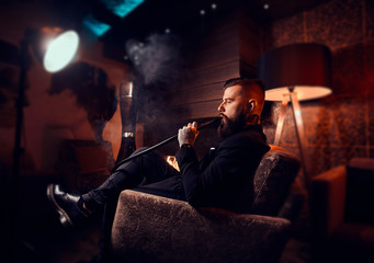 Fototapeta na wymiar Brutal tattooed man is making nice misty vapour while relaxing near fireplace and smoking hookah.