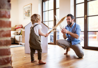 A toddler boy and father with carton swords playing indoors at home.