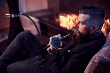 Attractive groomed man is relaxing on lounge near fireplace while smoking hookah. He has tattoo and watch on his hand.