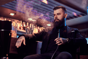 Photo of tattooed bearded man which is smoking hookah, making hazy vapour while chilling near bar counter.