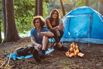 Attractive young couple have a rest in summer forest. People are sitting near tent and bonfire.