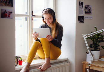 A young female student sitting on window sill, using tablet when studying.