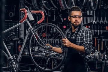 Fototapeta na wymiar Attractive pensive man in protective glasses is repairing bicycle at his own workshop.