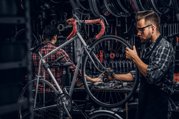 Obraz na płótnie Canvas Focused attractive man in glasses is chainging wheel for bicycle at busy workshop.