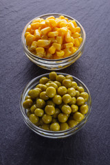 canned corn and green peas in a glass bowl on a dark stone background