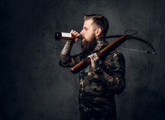 A bearded tattooed hipster guy in military shirt holding a medieval crossbow and drinks a craft beer. Studio photo against a dark wall
